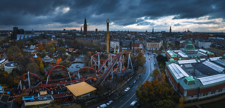 Aerial view of Tivoli Gardens' vibrant rollercoasters contrast with the muted cityscape under a dramatic sky, Copenhagen, Denmark.