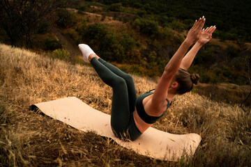 Female in Navasana variation on mat outdoors, core-focused yoga posture for abdominal strength, balance and fitness in scenic landscape, yoga boat pose in nature
