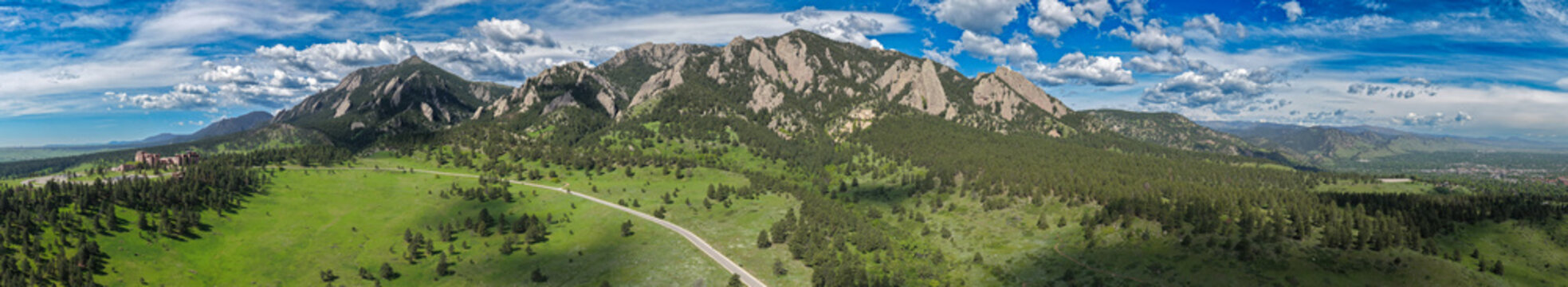 Aerial view of Boulder's Flatirons stand majestically against the sky, their rocky faces softened by lush greenery, a road winding through the landscape, Boulder, Colorado, United States.
