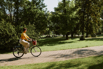 Fototapeta premium Middle aged Caucasian woman riding bicycle along paved path in green park, hands on handlebars, looking ahead, surrounded by tall trees and grass, enjoying outdoor activity