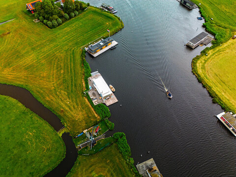 Aerial view of canal with moving boat, docks, windmill, and riverside buildings lush fields and water infrastructure blend tradition and recreation in a tranquil rural setting. - Powered by Adobe