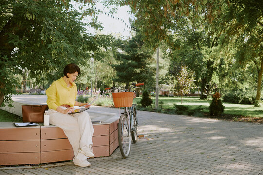Caucasian middle aged woman sitting on bench reading notebook outdoors in park, bicycle with basket parked nearby, brown leather bag and reusable cup placed beside her