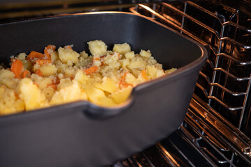 Black baking dish with sliced potatoes and carrots placed in a hot oven. Traditional Slovak meal preparation – French potatoes being baked at home.
