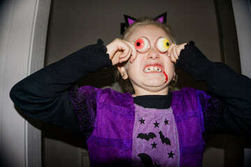 A young girl in a purple Halloween costume and cat ears playfully holds fake eyeballs to her eyes, with fake blood dripping from her mouth.