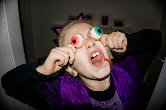 A young girl in a Halloween costume with fake eyeballs and blood splatters on her face makes a funny expression.