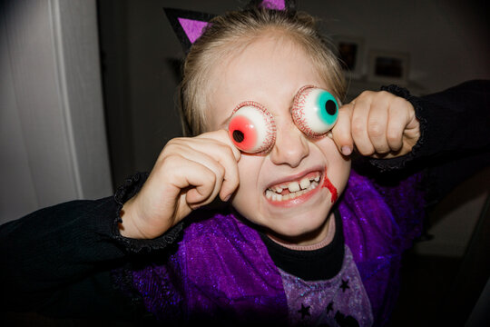 A young girl in a cat costume with fake blood on her face holds novelty eyeballs to her eyes, making a funny face.