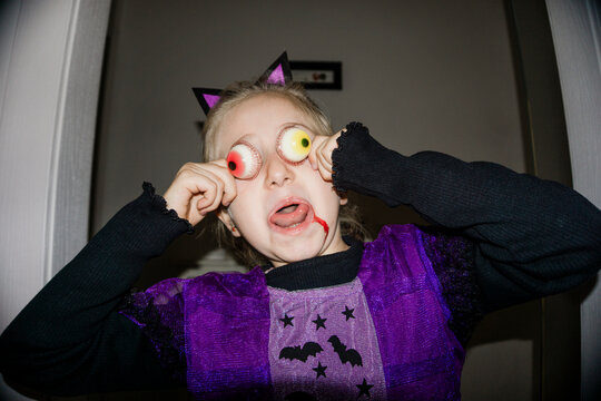 A young girl in a purple bat costume and cat ears playfully holds fake eyeballs to her eyes, with fake blood on her face.