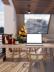 Blank screen laptop and coffee aside gift box on wooden counter table across Christmas tree in cafe.