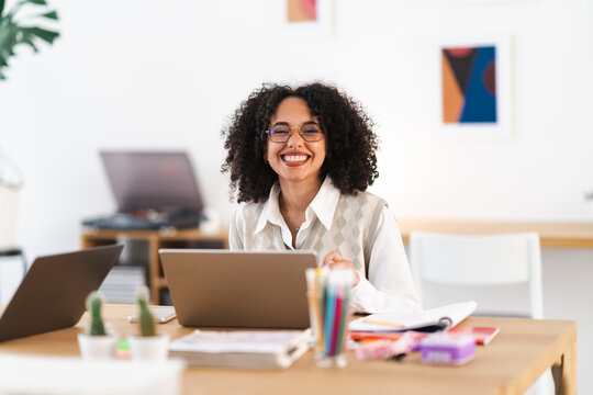 A young woman with curly hair smiles while working on her laptop at a desk filled with office supplies and plants.
