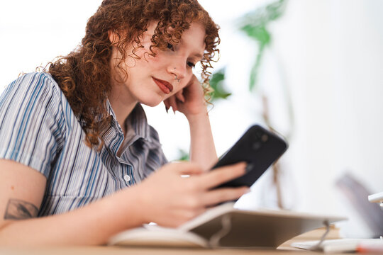 A young woman with curly red hair uses her smartphone while sitting at a desk with a laptop and notebook.
