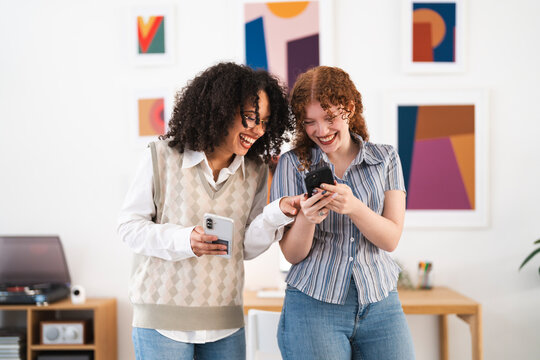 Two young women laugh together while looking at a smartphone in a bright, modern room with abstract art on the wall.
