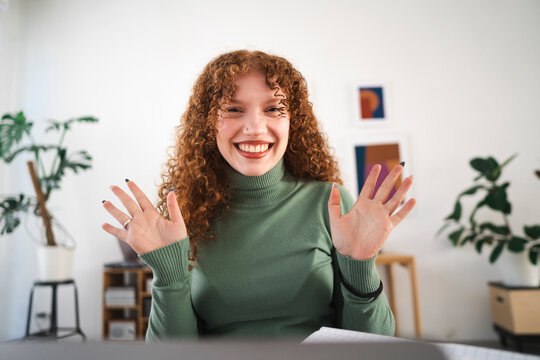 A young woman with curly red hair smiles and waves hello while working from home.