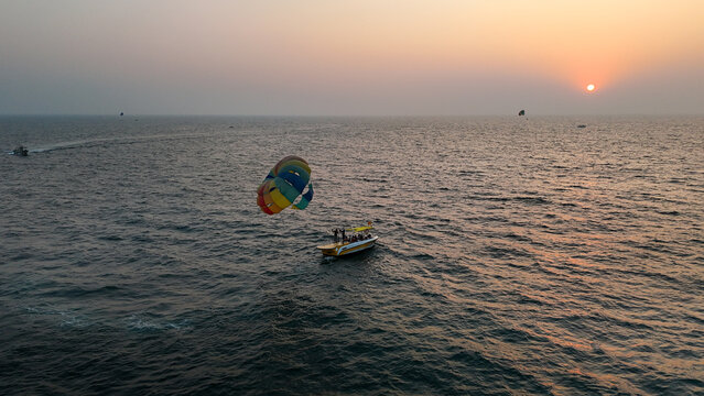 Aerial view of a vibrant parasail pulling a boat across the rippling, dark waters as the sun sets on the horizon, Goa, India.