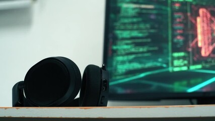 Black headphones sitting on a wooden shelf. In the blurry background, a computer monitor displays lines of scrolling green and red code