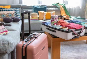 Suitcase packed with clothes and accessories for a summer vacation are placed next to a sofa in the living room, suggesting an upcoming trip. Open suitcase with colorful clothes ready for vacation.