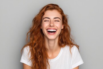 Against a plain background, a lively woman with fiery red locks and a white t-shirt is captured laughing with unbridled and genuine happiness