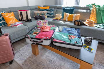 Open suitcase with summer clothes and accessories placed on coffee table and sofa in living room, symbolizing upcoming vacation and travel preparations