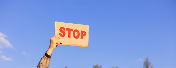 Raised hand holds a sign saying STOP against the backdrop of nature and blue sky.