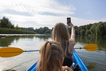 Mother taking a selfie with her daughter while kayaking on a calm lake surrounded by green trees on a sunny summer day. Happy family outdoor recreation and nature adventure