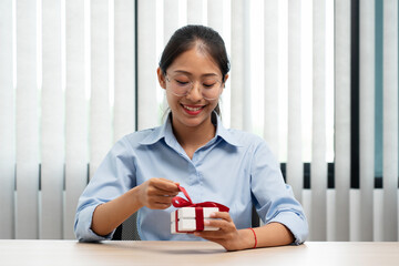 Happy woman is smiling and holding present box to wrapped with red ribbon while preparing gift for celebrate