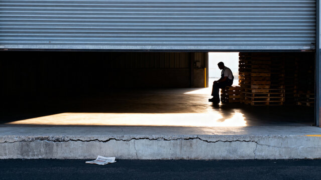 A lone worker sits silhouetted in the bright light of a partially open warehouse door, taking a quiet break in the surrounding darkness. - Powered by Adobe