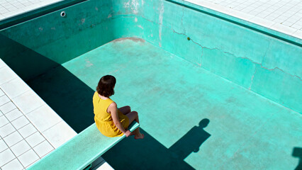 A lonely woman in a yellow dress sits on a diving board, contemplating a stark, empty swimming pool under the bright, harsh sun.