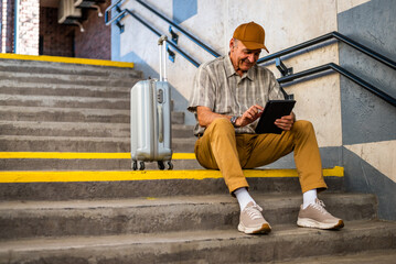 Happy senior man is sitting at railway station and waiting for boarding. He is using digital tablet. Senior people traveling.