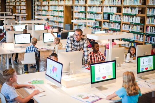 Children in library using computers for learning. Diverse group of kids engaged in educational activities. Teacher assisting students in a library setting. International students working on computers.
