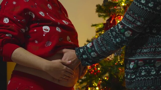 Medium shot of the hands of a pregnant couple in Christmas pyjamas trying to feel the baby kicks in front of the Christmas tree with lights