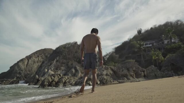 A low-angle, wide shot captures a man in swim shorts walking away from the camera and along the water's edge on a sandy beach.