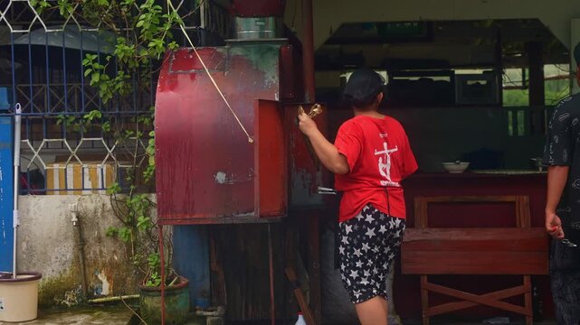 A still shot of street BBQ vendor in red shirt and star‑patterned pants, fanning heat over red metal grill outside their store along the road of Lucban, Quezon Province Philippines