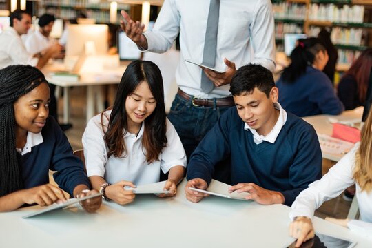 Diverse group of young students studying together, using tablets in a library. Students collaborating, studying, and using technology. Group of students in library setting. High school education.