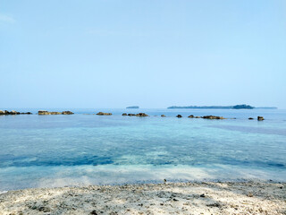 View of beautiful beach coast line with sea rocks. Clear open water ocean with amazing blue color and white sand, under blue sunny sky. Nature beach and sea photography.