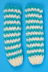 An overhead studio shot displays a pair of chunky knitted warmers in a teal and cream two-color checkerboard pattern on a vibrant blue background