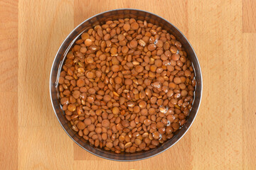 An overhead shot shows a stainless steel bowl filled with brown lentils soaking in water, placed on a light wooden background