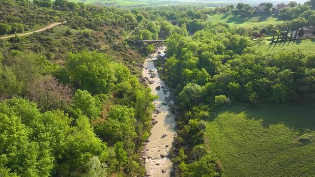 Aerial view of a bridge spanning a river, surrounded by vibrant green trees and rolling hills, creating a scene of natural beauty, Bagno Vignoni, Tuscany, Italy.