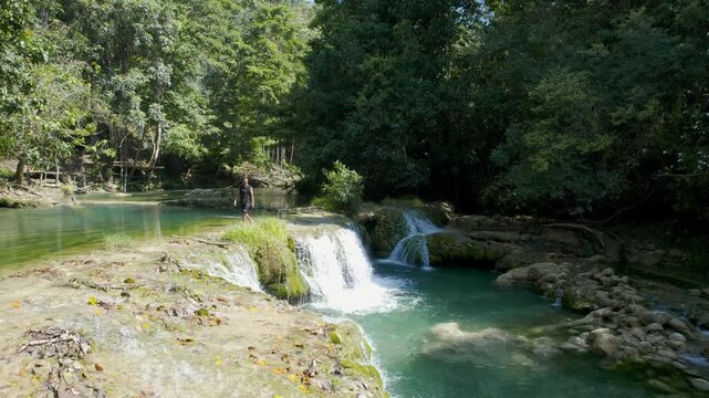 A traveler walks along the edge of Cunca Rami Waterfall in West Manggarai, Flores. The turquoise water cascades over mossy rocks surrounded by lush tropical forest, creating a serene natural scene.

