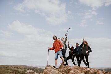 Group of diverse friends hiking on a rocky hilltop, holding trekking poles, enjoying nature and adventure under a cloudy sky, celebrating friendship and exploration. Happy friends on a hike.