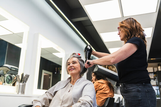 Professional hairstylist drying a happy senior woman's hair in a modern hair salon, providing beauty and pampering service