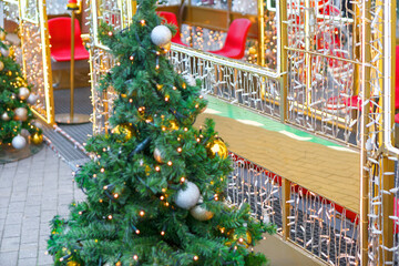 Glowing Christmas tree with silver ornaments stands amid twinkling white lights and red chairs, creating a joyful, cozy holiday scene. Reflections on brick pavement enhance the festive ambiance