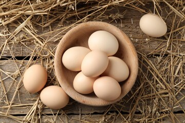 Raw chicken eggs in bowl and straw on wooden table, flat lay