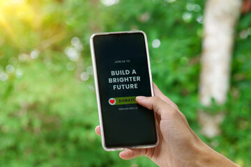 Hand holding smartphone showing donate message build a brighter future concept with green nature background