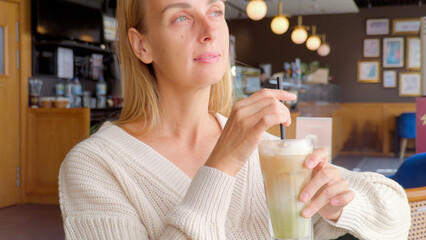 Woman enjoying iced matcha latte in cozy cafe wearing white sweater and relaxing alone