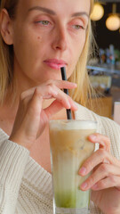 Woman stirring iced matcha latte with straw while sitting in cafe wearing white knitted sweater