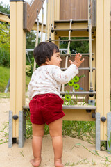 Obraz premium First steps of a cute mixed-race, Latina, and Asian girl with dark hair playing happily in a playground, trying to climb the stairs of a swing while waving her hand.
