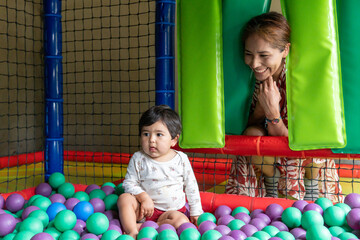 A happy Latina mother plays with her multiracial little boy in a multicolored ball pit.