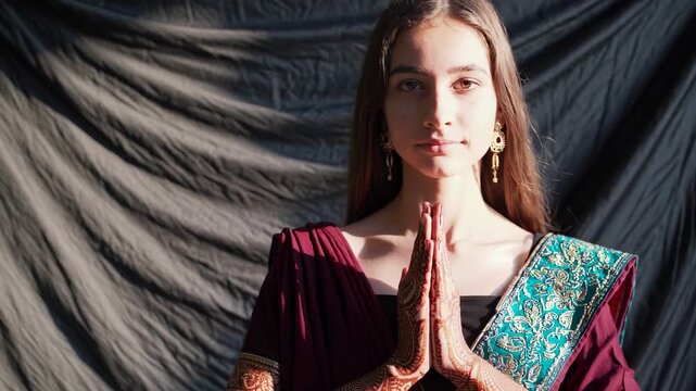 Woman performing namaste greeting and prayer. Portrait in saree with henna on hands. Traditional prayer pose conveys meditative respect and calm. Closeup portrait shows embroidered pallu and jewelry.