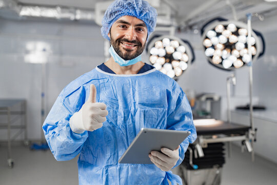 A smiling surgeon in scrubs gives a thumbs up while holding a tablet in an operating room. - Powered by Adobe
