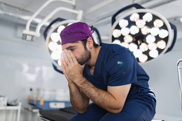 A stressed surgeon sits in an operating room, covering his face with his hands, illuminated by...