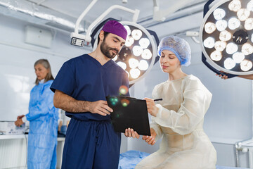 A surgeon reviews paperwork with a patient in an operating room, with another medical professional in the background.